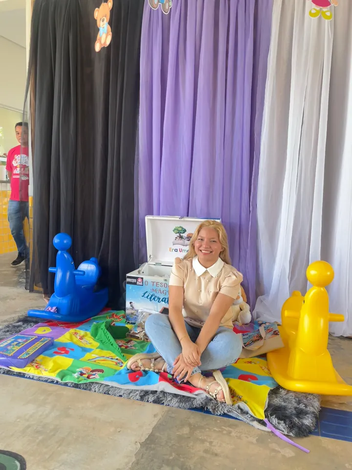 Lanna Campos sorrindo, sentada no chão entre brinquedos coloridos, em frente a uma cortina lilás.