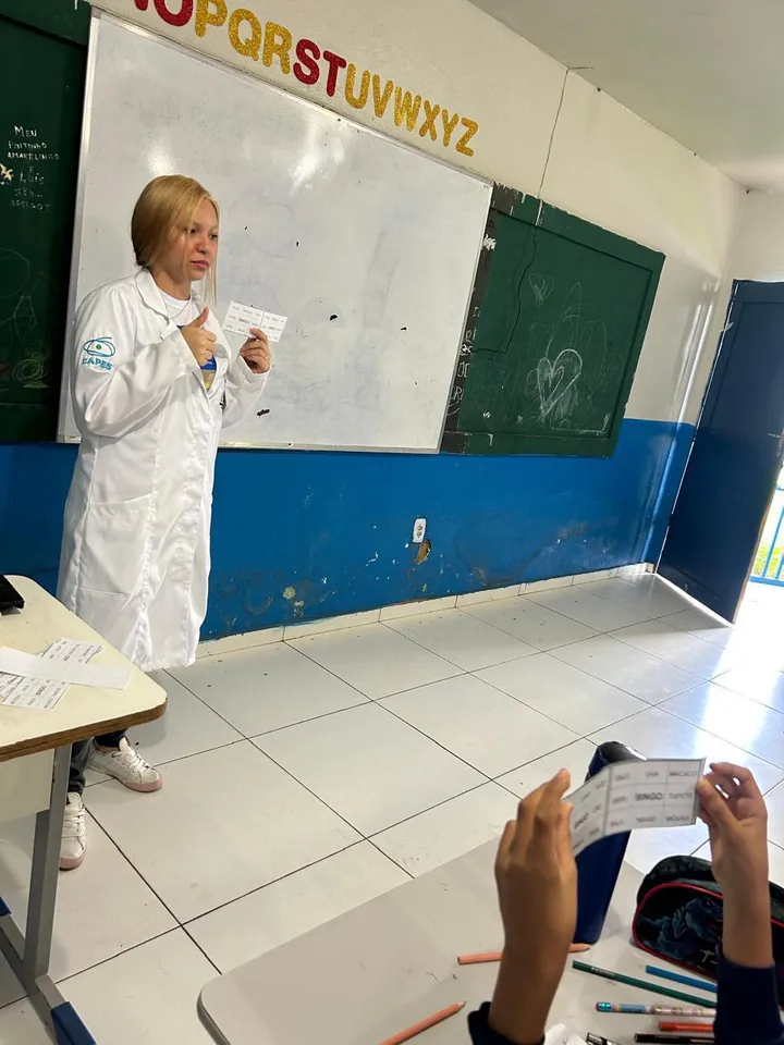 Lanna Campos de jaleco em sala de aula, lendo diante de crianças sentadas nas carteiras.