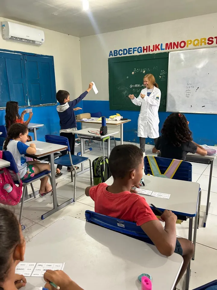 Sala de aula com crianças em atividade e Lanna Campos de jaleco ao fundo.