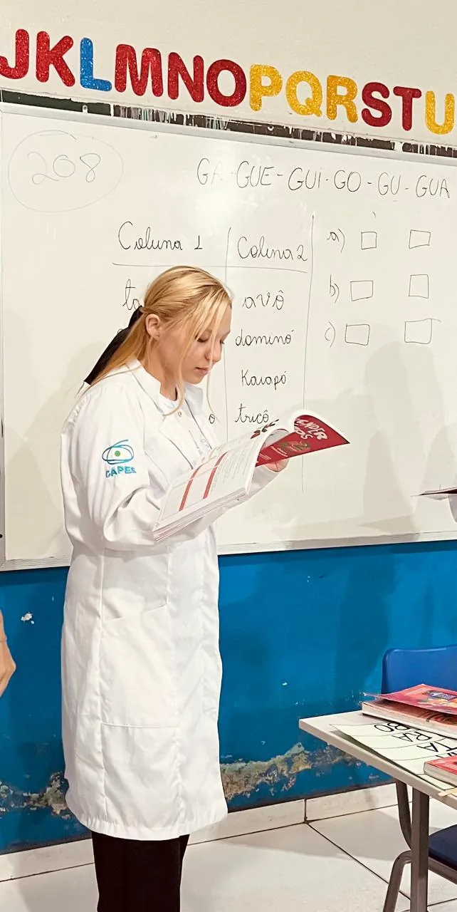 Lanna Campos de jaleco lendo um livro diante do quadro branco de uma sala de aula.
