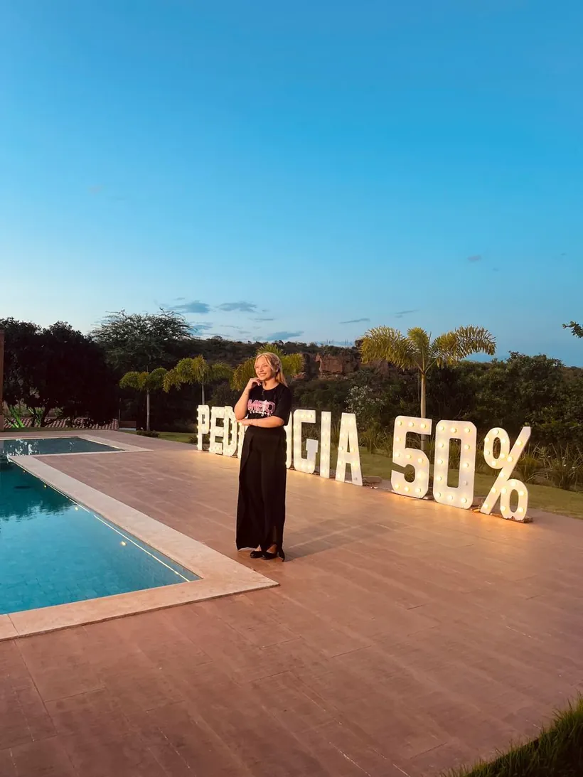 Lanna Campos de vestido preto ao lado das letras luminosas Pedagogia 50%, à beira de uma piscina.