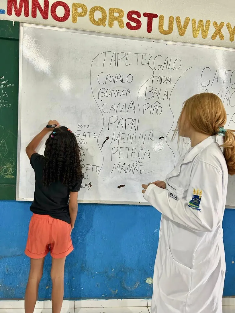 Lanna Campos, de jaleco, ao lado de uma criança que escreve palavras no quadro de uma sala de aula.