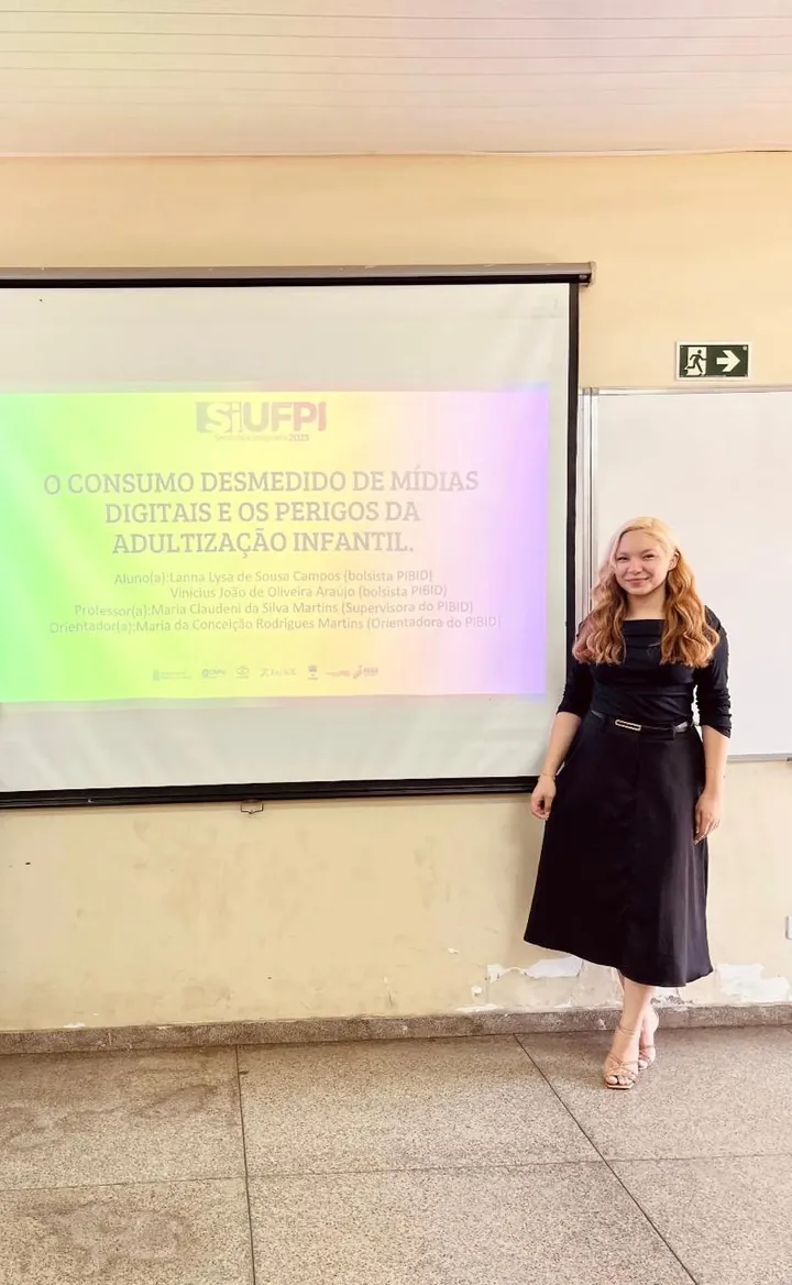 Lanna Campos ao lado de um slide sobre o consumo desmedido de mídias digitais, em apresentação na UFPI.